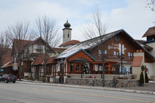 A charming Bavarian-style building with a clock tower, surrounded by trees and snow, along a quiet street.