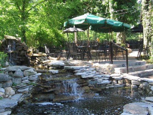 A serene outdoor setting featuring a stone waterfall, a pond, and a dining area with green umbrellas surrounded by lush greenery.