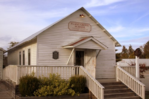 Historic Flagg Grove School building with a wooden exterior and a sign, surrounded by greenery and a clear sky.