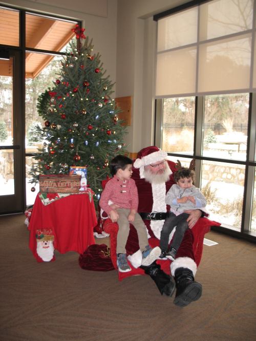 Two children sit on Santa's lap in a festive room with a Christmas tree and decorations.