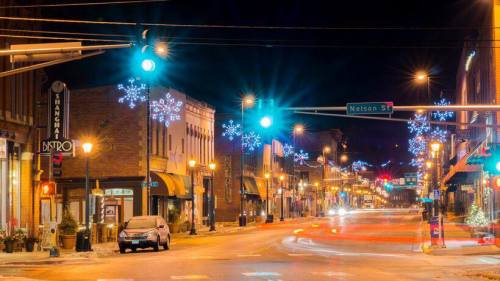 A quiet street at night, adorned with snowflake lights, with traffic signals and storefronts illuminated.