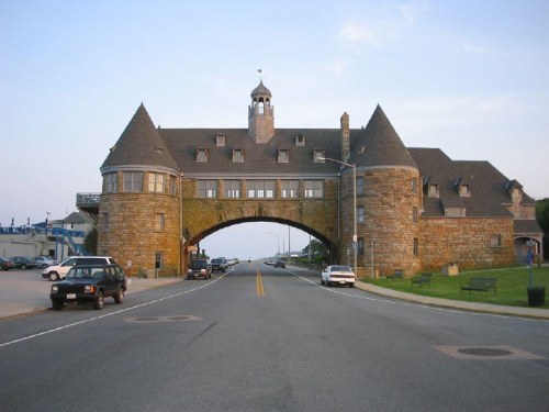 Historic stone building with towers and an archway, viewed from a road lined with parked cars and benches.