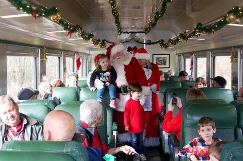 Santa and Mrs. Claus greet children on a festive train, surrounded by families in a decorated car.