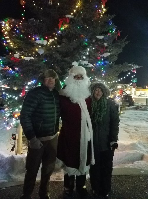 A couple poses with Santa in front of a brightly lit Christmas tree at night, surrounded by snow.