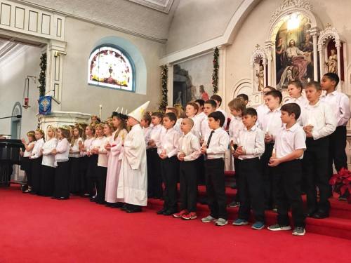 A group of children in white shirts stands on a red carpet in a church, performing in front of a decorated altar.