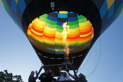 A colorful hot air balloon inflates as people wave from the basket, with flames visible from the burner below.