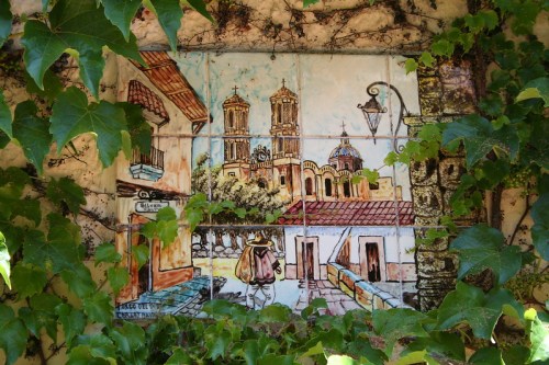 A colorful mural depicting a village scene with towers, a street, and a person walking, framed by green ivy.