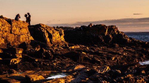 Silhouetted figures on rocky coastline at sunset, with golden light illuminating the rocks and ocean in the background.