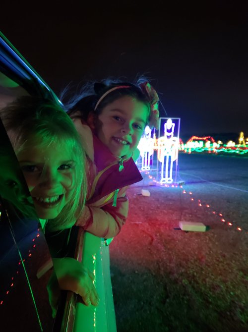Two smiling girls lean out of a car window, surrounded by colorful holiday lights in the background.