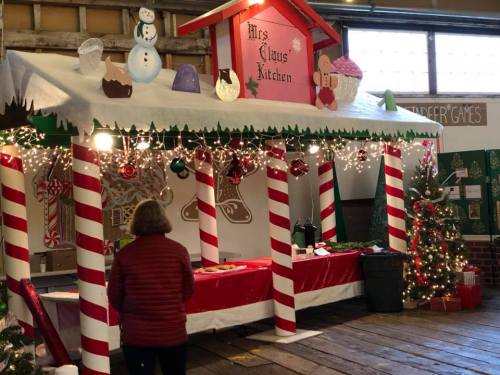A festive holiday booth decorated with candy canes, lights, and a sign reading "Mrs. Claus Kitchen," with a person standing nearby.