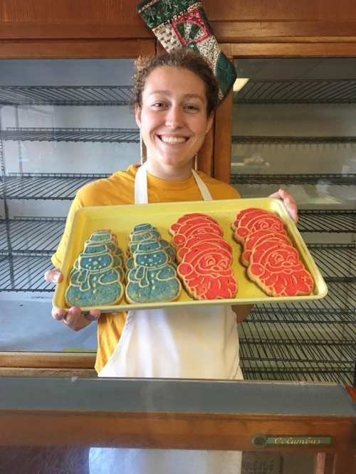 A smiling person holds a tray of decorated cookies, featuring blue and red designs, in a bakery setting.
