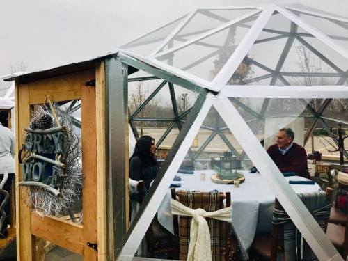 Two people sit at a table inside a transparent geodesic dome, surrounded by winter decor and a gray wolf sign.