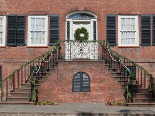 A brick building with black shutters, a festive wreath on the door, and garlands draped along the staircase railing.