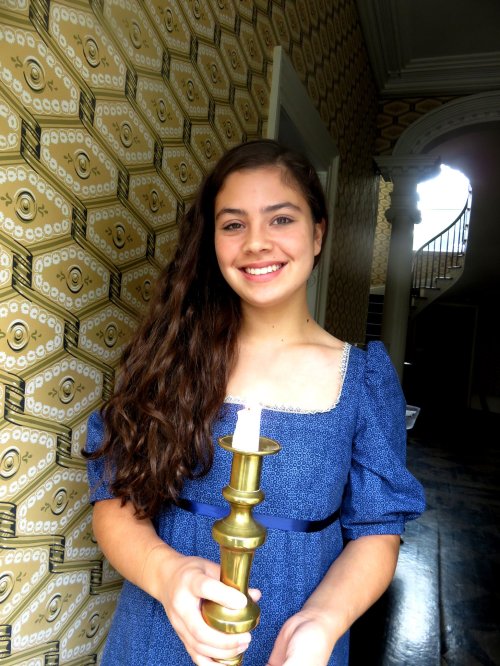A young girl in a blue dress holds a brass candlestick, smiling in a vintage-style room with patterned wallpaper.