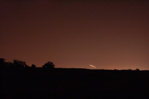 A dark landscape silhouette against a starry night sky, with a faint light streaking across the horizon.