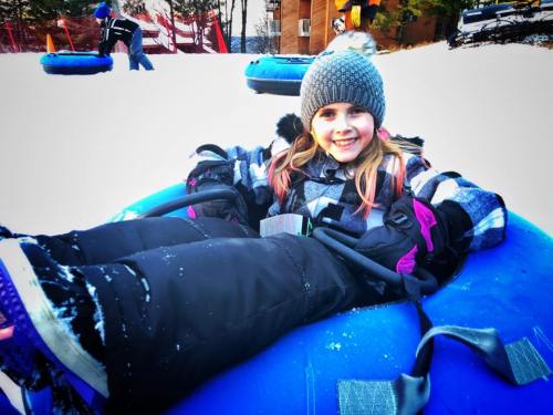 A smiling girl in winter attire sits in a blue snow tube, ready to slide down a snowy hill.
