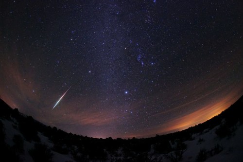 A starry night sky with a bright meteor streaking across, surrounded by constellations and a faint horizon glow.