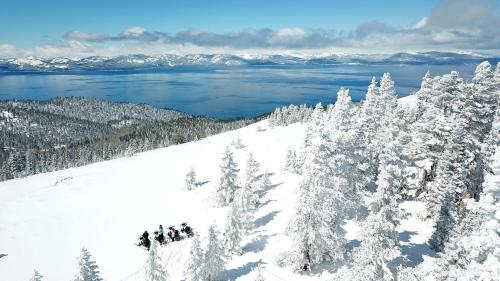 A snowy landscape with evergreen trees, overlooking a lake and mountains under a clear blue sky.