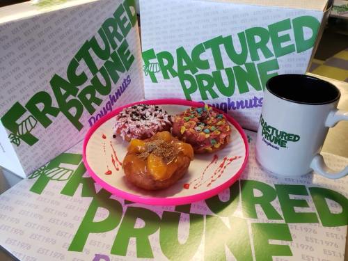 A plate with colorful donuts sits in front of branded boxes and a mug from Fractured Prune Doughnuts.