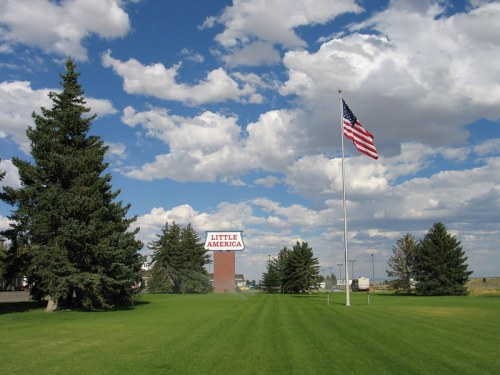 A grassy area with a large sign reading "Little America" and an American flag waving under a blue sky with clouds.