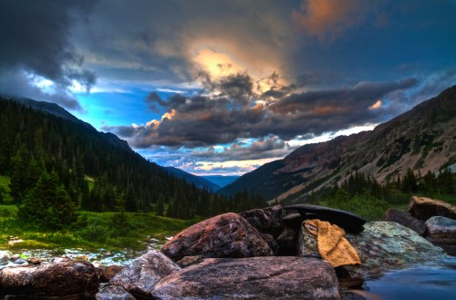 A serene mountain landscape with rocky foreground, lush greenery, and dramatic clouds in a colorful sky.