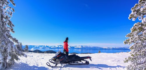 A person in a red jacket stands beside a snowmobile, overlooking a snowy landscape and a blue lake under a clear sky.