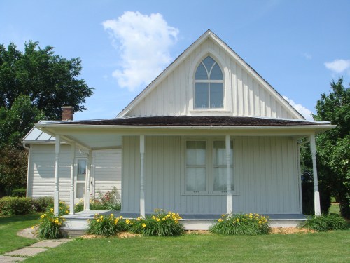 A white, gabled house with a front porch and yellow flowers in the garden, set against a blue sky with clouds.