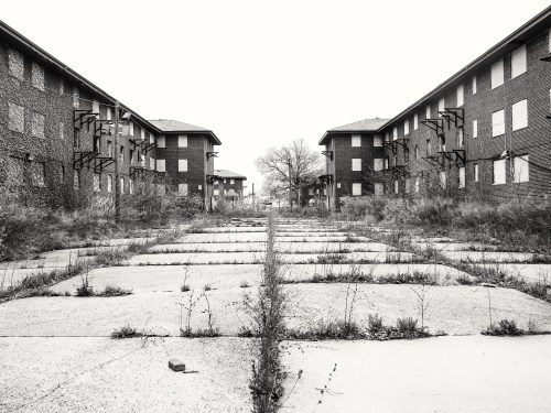Abandoned buildings flanking a cracked concrete path, overgrown with weeds, under a gray sky.