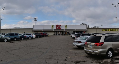 A Kmart store with a large parking lot, featuring various parked cars and a cloudy sky above. Store closing sign visible.