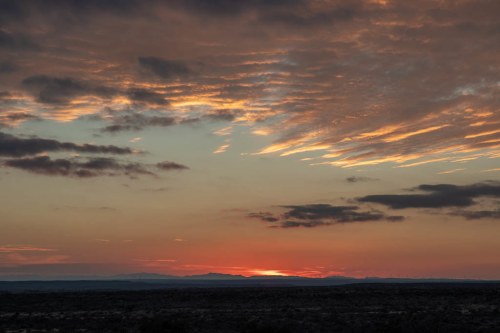 A vibrant sunset over a vast landscape, with colorful clouds and distant mountains silhouetted against the horizon.