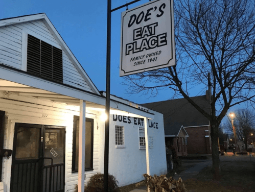 A family-owned restaurant, Doe's Eat Place, with a sign and a cozy exterior, illuminated at dusk.