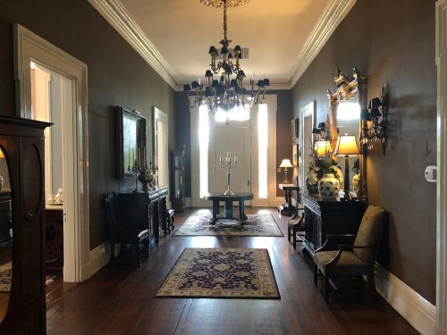 A spacious hallway with dark walls, elegant furniture, a chandelier, and decorative rugs, leading to a bright entrance.