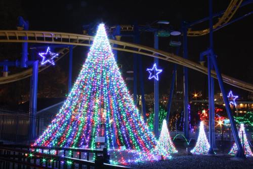 A brightly lit Christmas tree and stars illuminate a festive scene near a roller coaster at night.