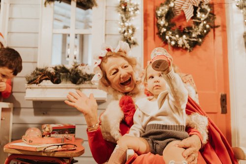 A joyful elderly woman waves while holding a toddler who is showing a toy, festive decorations in the background.