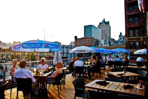 A rooftop restaurant with diners enjoying meals under blue umbrellas, overlooking a city skyline at sunset.
