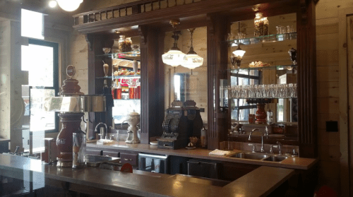 A vintage-style bar with wooden shelves, glassware, and retro decor, featuring a soda fountain and cash register.