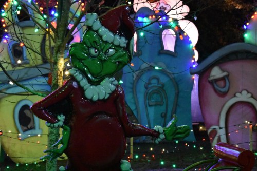 A festive Grinch statue in a Santa outfit, surrounded by colorful holiday lights and whimsical, decorated houses.