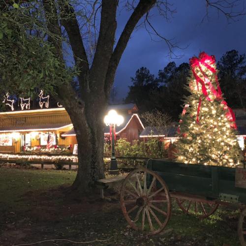 A festive scene featuring a decorated Christmas tree, a wagon, and holiday lights on buildings under a twilight sky.
