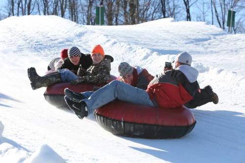 Four people in winter clothing sliding down a snowy hill on inner tubes, enjoying a fun day of tubing.