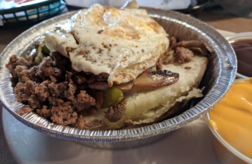 A metal dish filled with a baked potato topped with ground meat, sautéed vegetables, and a fried egg, with sauces on the side.