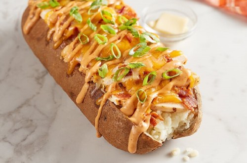 Baked potato topped with cheese, bacon, green onions, and a drizzle of sauce on a marble surface.