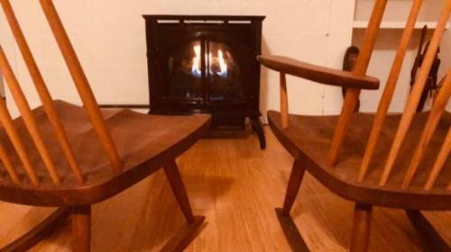 Two wooden rocking chairs facing a small fireplace with a warm flame, set in a cozy indoor space.