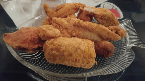 A glass plate filled with various fried foods, including chicken, fish, and corn fritters.