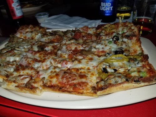 A large pizza topped with cheese, pepperoni, vegetables, and herbs, served on a white plate in a dimly lit setting.