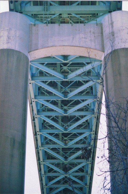 View from below a bridge, showcasing its blue steel structure and concrete pillars against a clear sky.