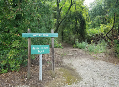 A wooden sign marks the entrance to the Jesse James Trail, surrounded by lush greenery and a gravel path.