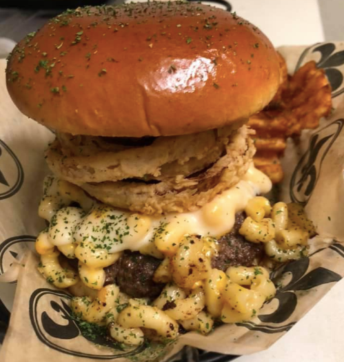 A gourmet burger topped with macaroni and cheese, crispy onion rings, and served with curly fries.