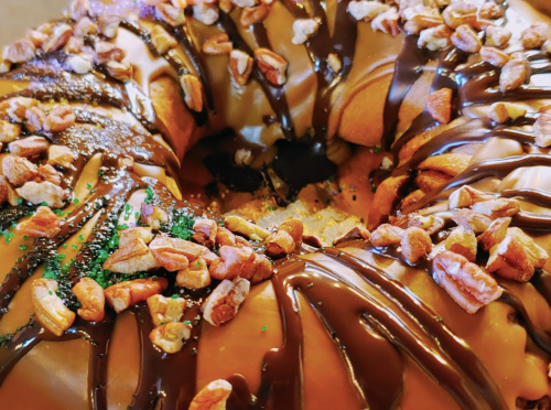 A close-up of a chocolate-drizzled donut topped with pecans and green sprinkles.