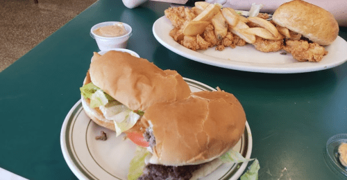 Two burgers with lettuce and tomato on a green table, alongside a plate of fried chicken and fries.