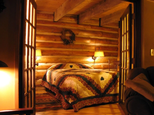 Cozy log cabin bedroom with a quilted bed, warm lighting, and wooden walls, creating a rustic atmosphere.